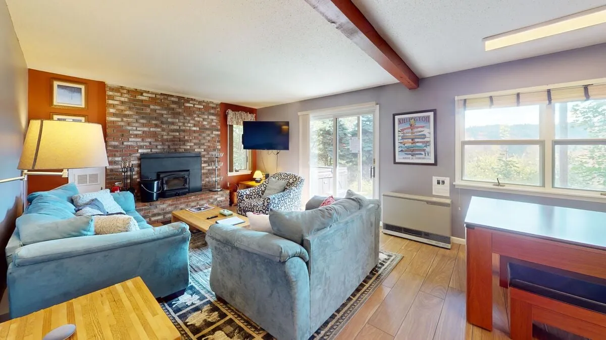 A cozy living space with a brick fireplace, plush sofas, large windows, and a wooden table. The room is arranged for both relaxation and productivity, providing ample room for socializing or working by the fire.