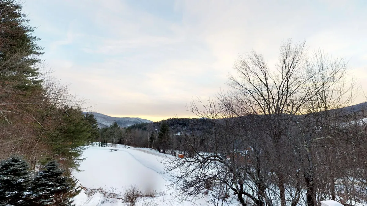 A tranquil winter landscape unfolds from the property, with snow-covered ground, bare trees, and distant mountains under a clear sky. The scene evokes the serenity and natural beauty of the Vermont region, inviting guests to step out and explore.