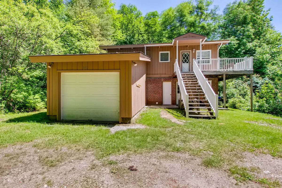 This exterior image shows the front entrance of the chalet surrounded by lush green foliage and a welcoming porch. The scene evokes a sense of summertime tranquility and highlights the property's connection to the outdoors. The visible stairs and garage door indicate accessibility and ample space for outdoor gear.