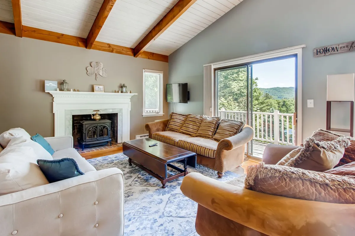 A cozy living room with a crackling fireplace, large windows revealing mountain views, plush sofas, and a coffee table. The abundant natural light and inviting seating area make it an ideal space for gathering, storytelling, or unwinding after outdoor adventures.
