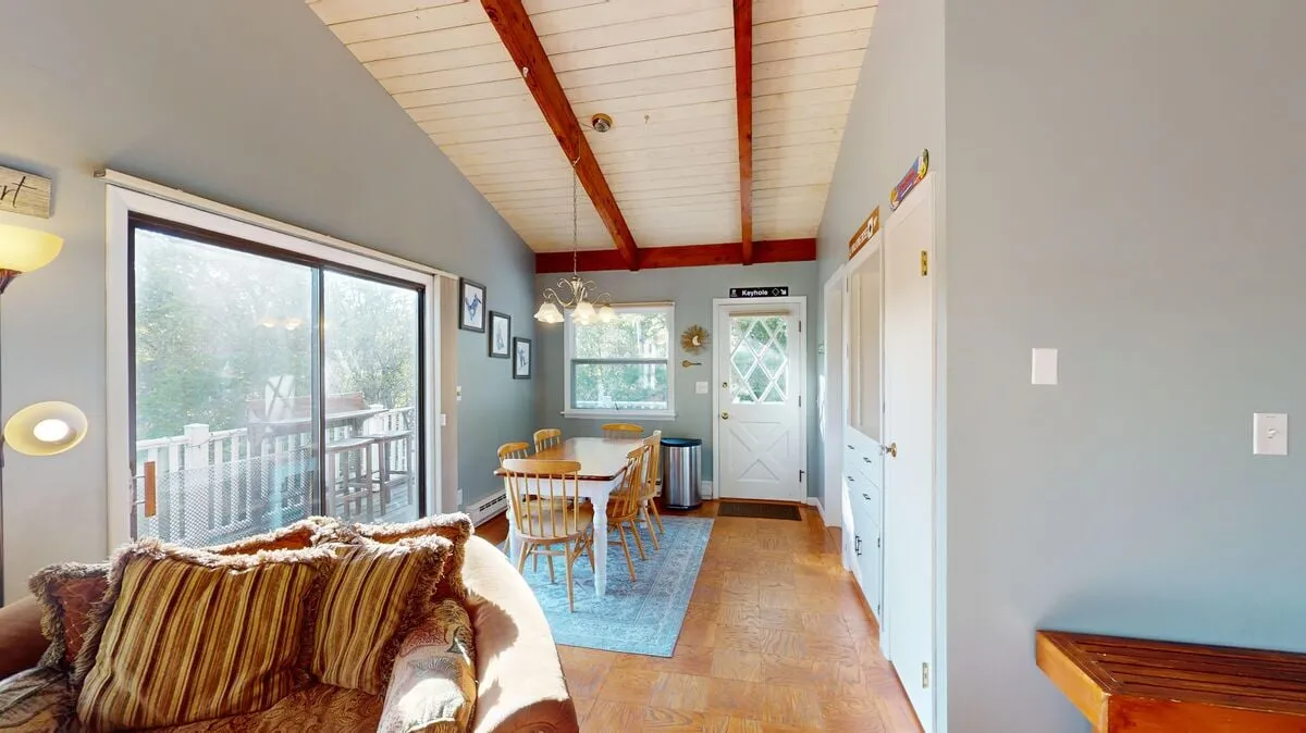 A bright, inviting open-plan area showcasing a comfortable sofa, a large dining table, and expansive windows letting in natural light. The arrangement is ideal for group gatherings, meals, or settling in with a laptop for remote work.