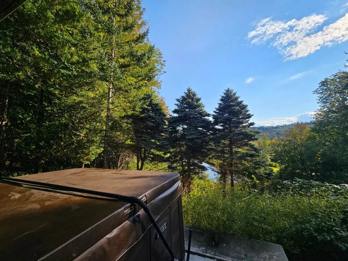 A steaming outdoor hot tub set amid trees, with a serene water view in the background. The natural surroundings and inviting hot tub evoke relaxation and comfort, ideal for soothing tired muscles after skiing or snowmobiling.